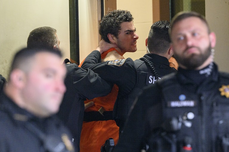Luigi Mangione wearing an orange jumpsuit makes an angry face as police officers push him, one with a hand on his neck