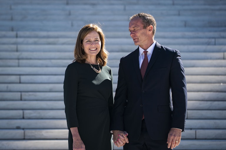 Amy Coney Barrett smiles and holds hands with her husband, who looks at her. In the background are the steps in front of the Supreme Court building.