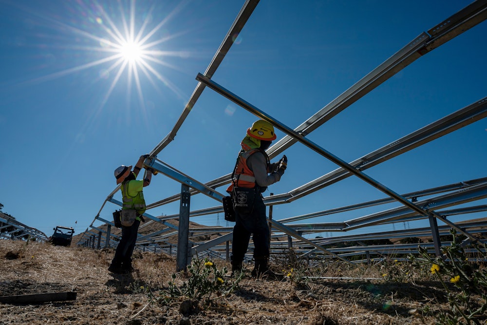 This image taken from soil level looks up at two workers wearing helmets and reflective gear holding power tools to skeletal metal frames outdoors against the bright sun.