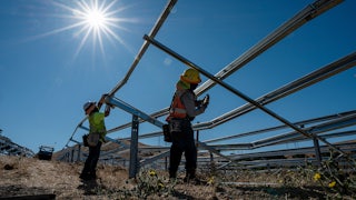 This image taken from soil level looks up at two workers wearing helmets and reflective gear holding power tools to skeletal metal frames outdoors against the bright sun.