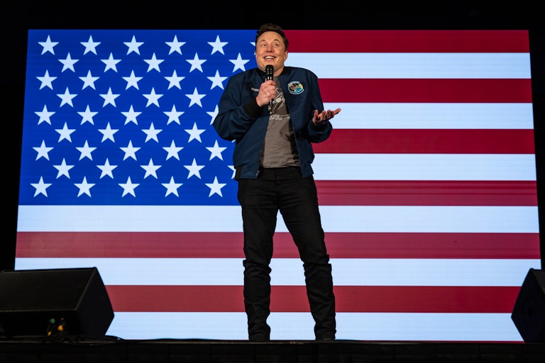 Elon Musk stands in front of an American flag backdrop holding a microphone in his right hand with his left hand making a shrugging motion.