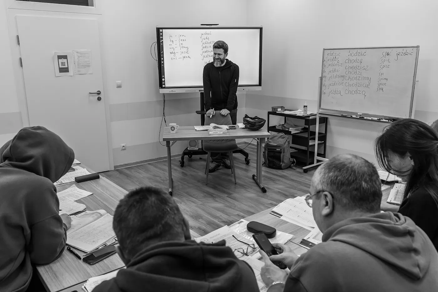 A black and white photograph of Michal Golubiewski, a language teacher, teaching Polish to Ukrainian refugees at Warsaw’s Jesuit refugee housing center.