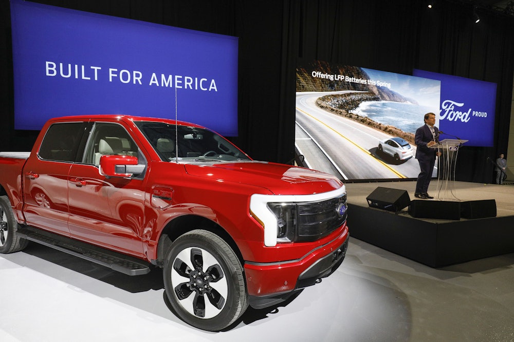 A man stands on a small stage next to a massive, red pickup truck with a screen behind it stating "Built for America."