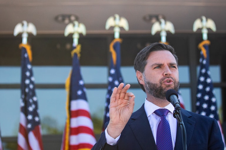 J.D. Vance speaks in front of a mic. A row of U.S. flags is behind him.