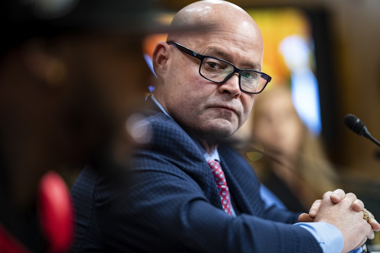A bald man wearing a suit crosses his hands while sitting at a table.