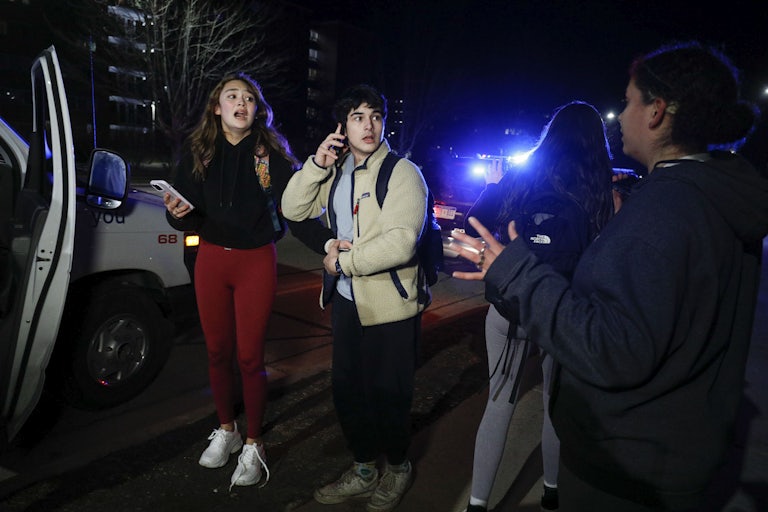 A group of three four students stands outside at night. Two of them are on the phone and look afraid.