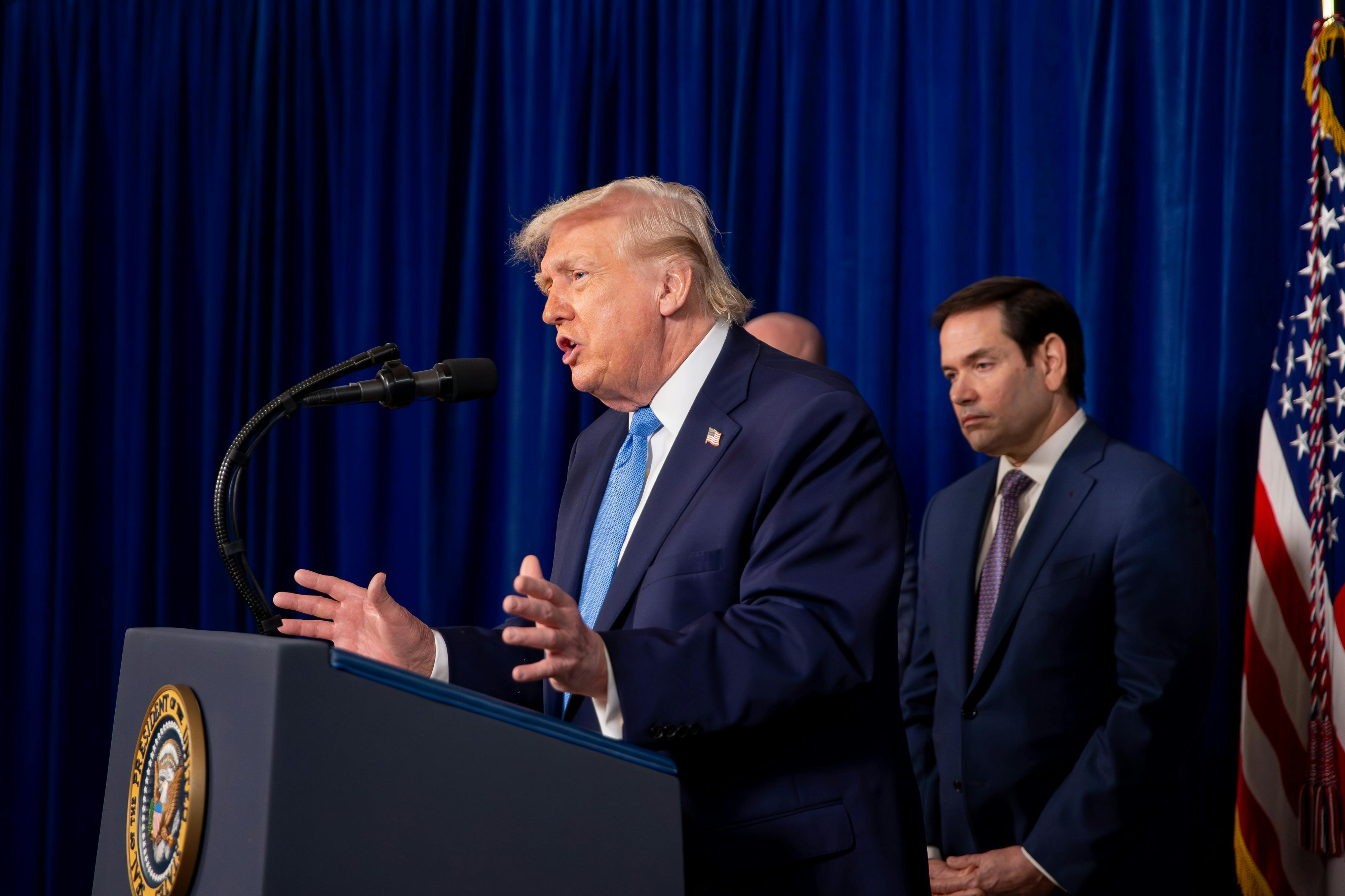 Donald Trump gestures and speaks at a podium while Secretary of State Marco Rubio stands behind him