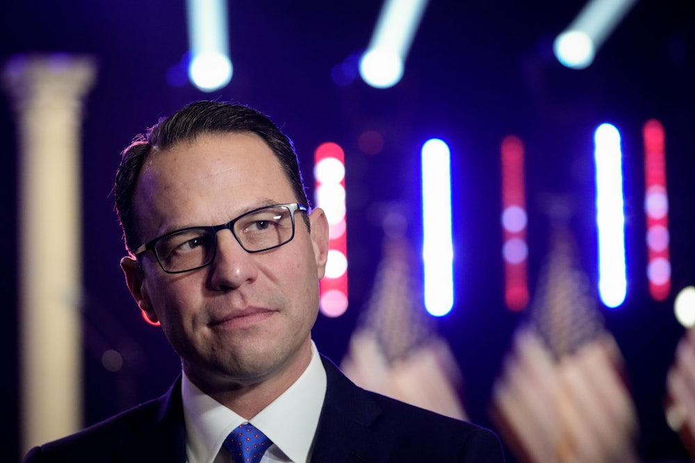 Josh Shapiro, wearing glasses, a suit, and a blue tie, looks quizzically as three American flags stand in the background.