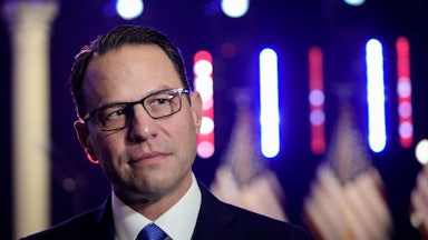 Josh Shapiro, wearing glasses, a suit, and a blue tie, looks quizzically as three American flags stand in the background.