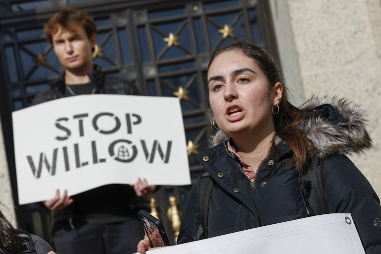 A student speaks with another, behind, holding a sign saying "Stop Willow."