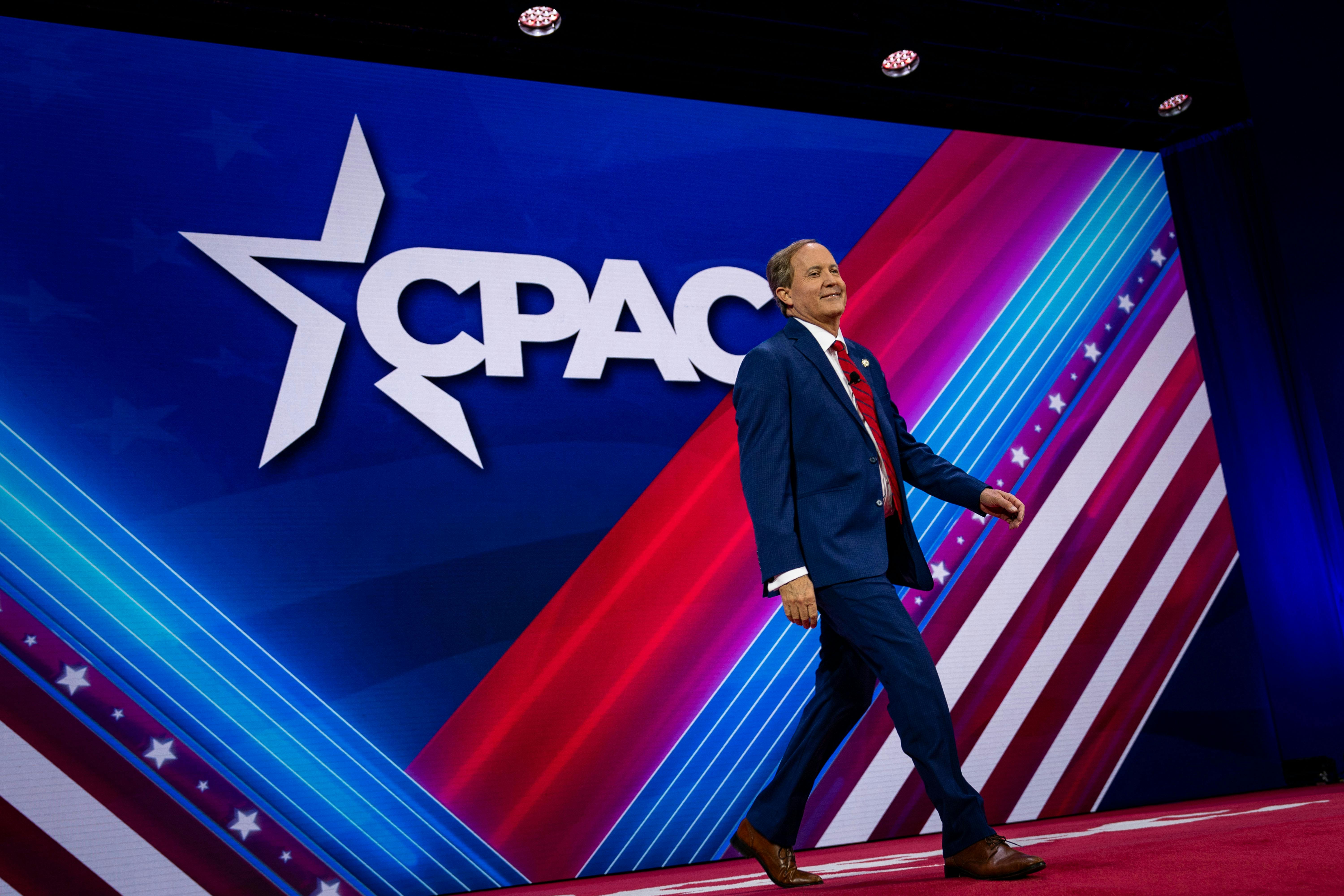 Ken Paxton walks across a red, white, and blue stage with the CPAC logo behind him.