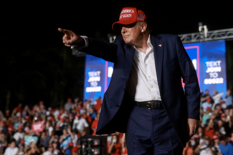 Donald Trump wearing a red MAGA cap smiles and points to the crowd at one of his rallies
