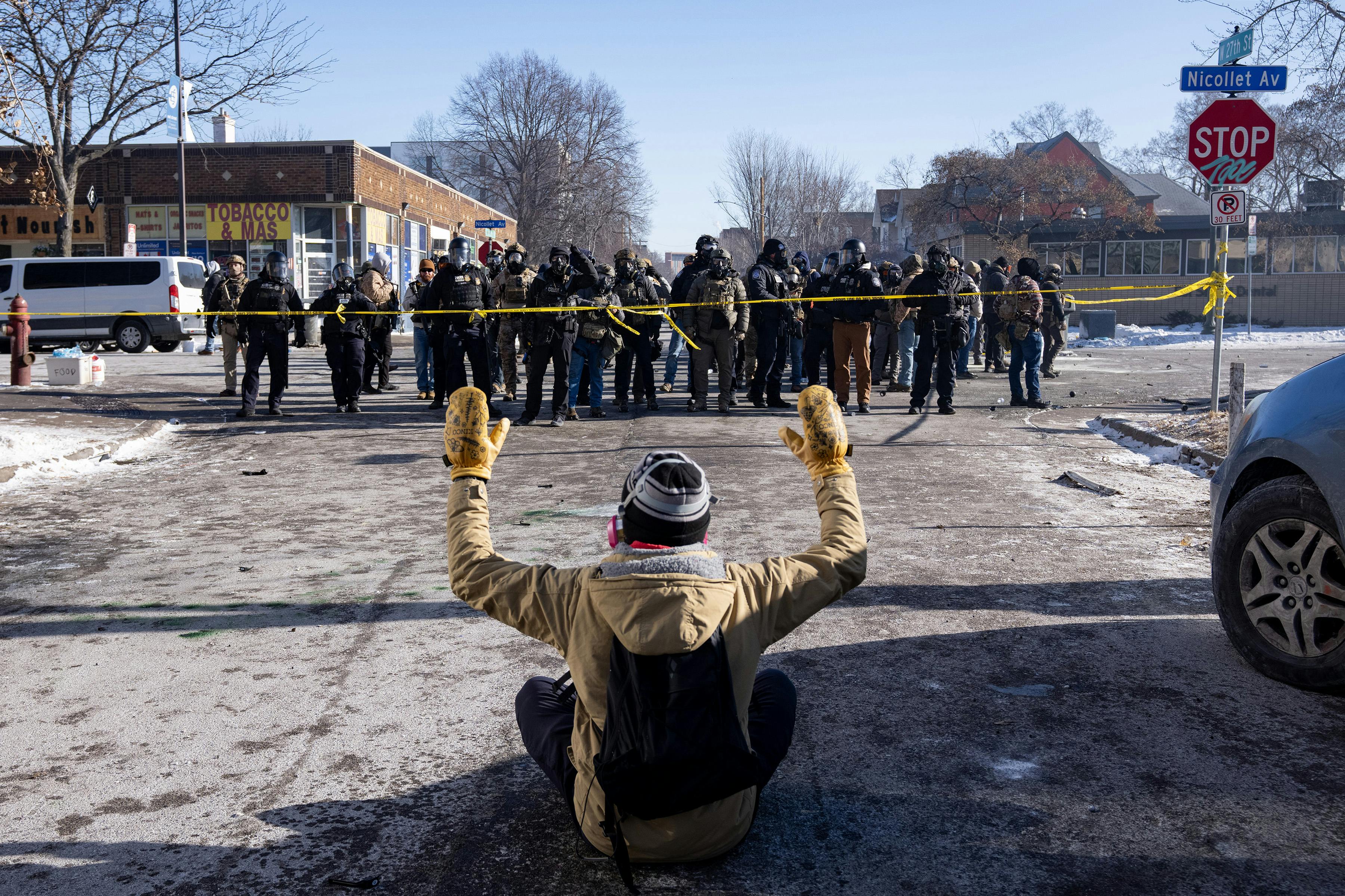 A person sits on the ground with their hands above their head as a group of masked federal agents walk towards them