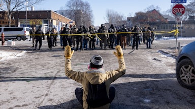 A person sits on the ground with their hands above their head as a group of masked federal agents walk towards them