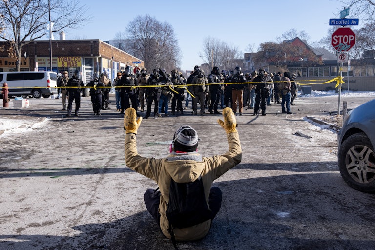 A person sits on the ground with their hands above their head as a group of masked federal agents walk towards them