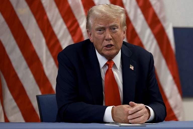 Donald Trump sits at a table. A large U.S. flag is behind him.