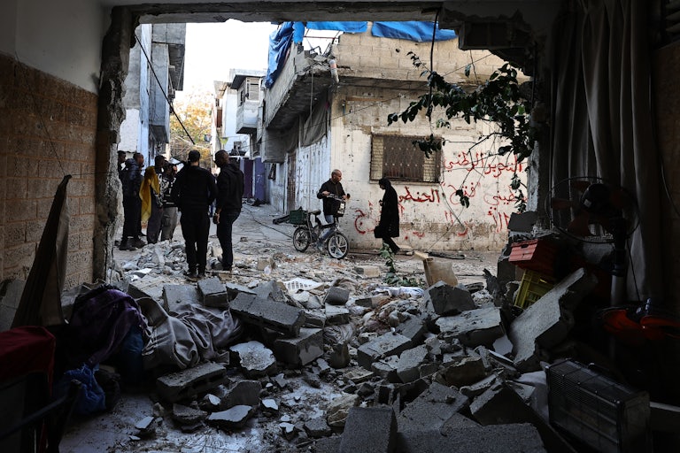 A wall to a building is completely torn off, rubble everywhere, as people stand outside talking to each other.