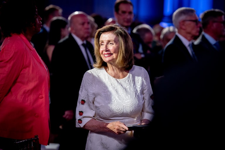 Nancy Pelosi smiles, dressed in formal evening wear. Others are in the background.