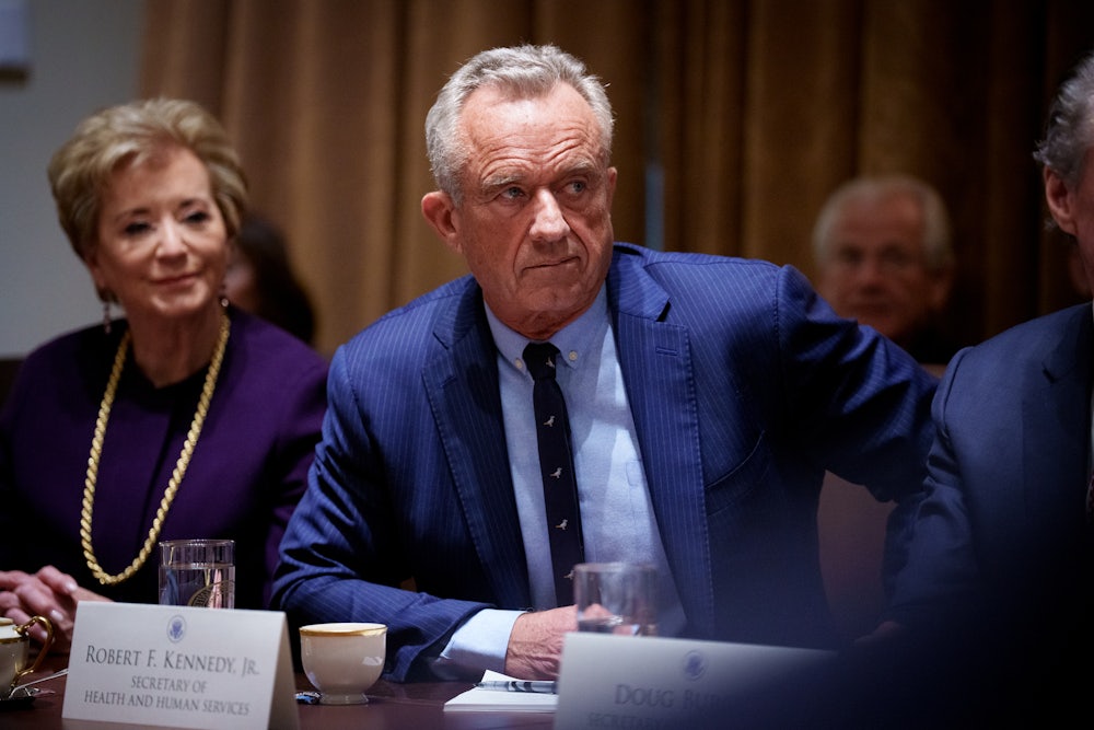 RFK Jr. wears a blue suit while seated at a table next to Linda McMahon, with a name card in front of him.