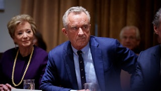 RFK Jr. wears a blue suit while seated at a table next to Linda McMahon, with a name card in front of him.