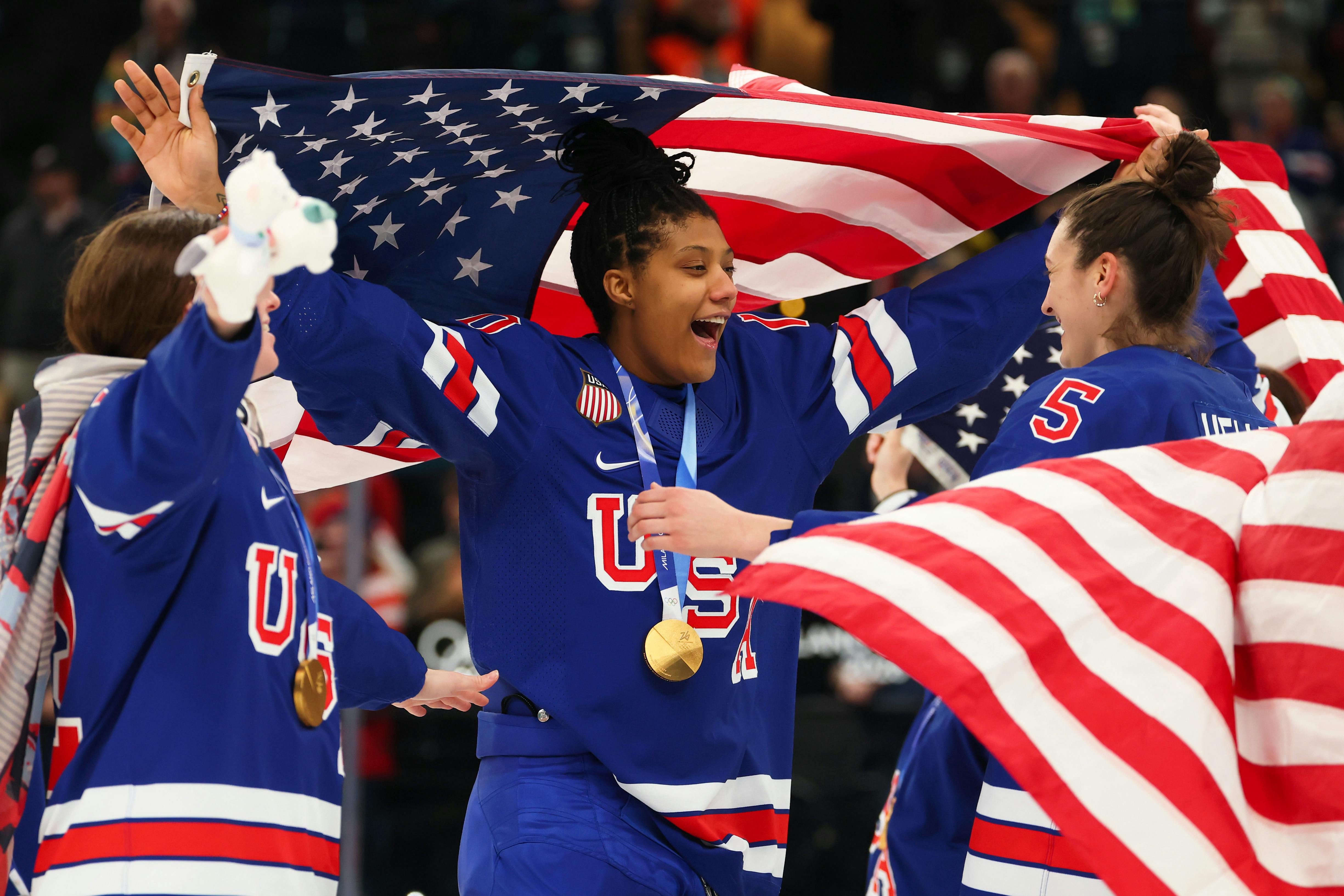 Three Team USA women hockey players celebrate on the rink with large U.S. flags.