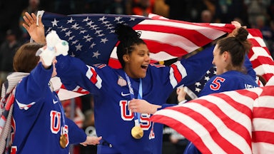 Three Team USA women hockey players celebrate on the rink with large U.S. flags.