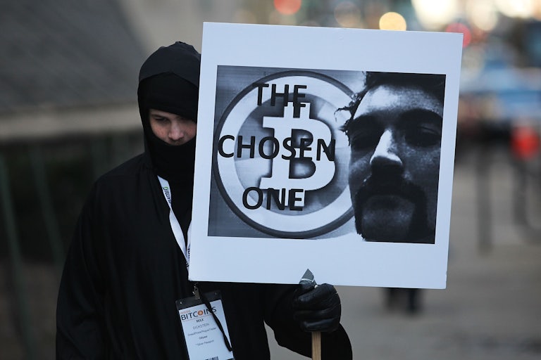 A man dressed in all black and a balaclava holds a sign that says "The Chosen One" with the Bitcoin logo and a photo of Ross Ulbricht.