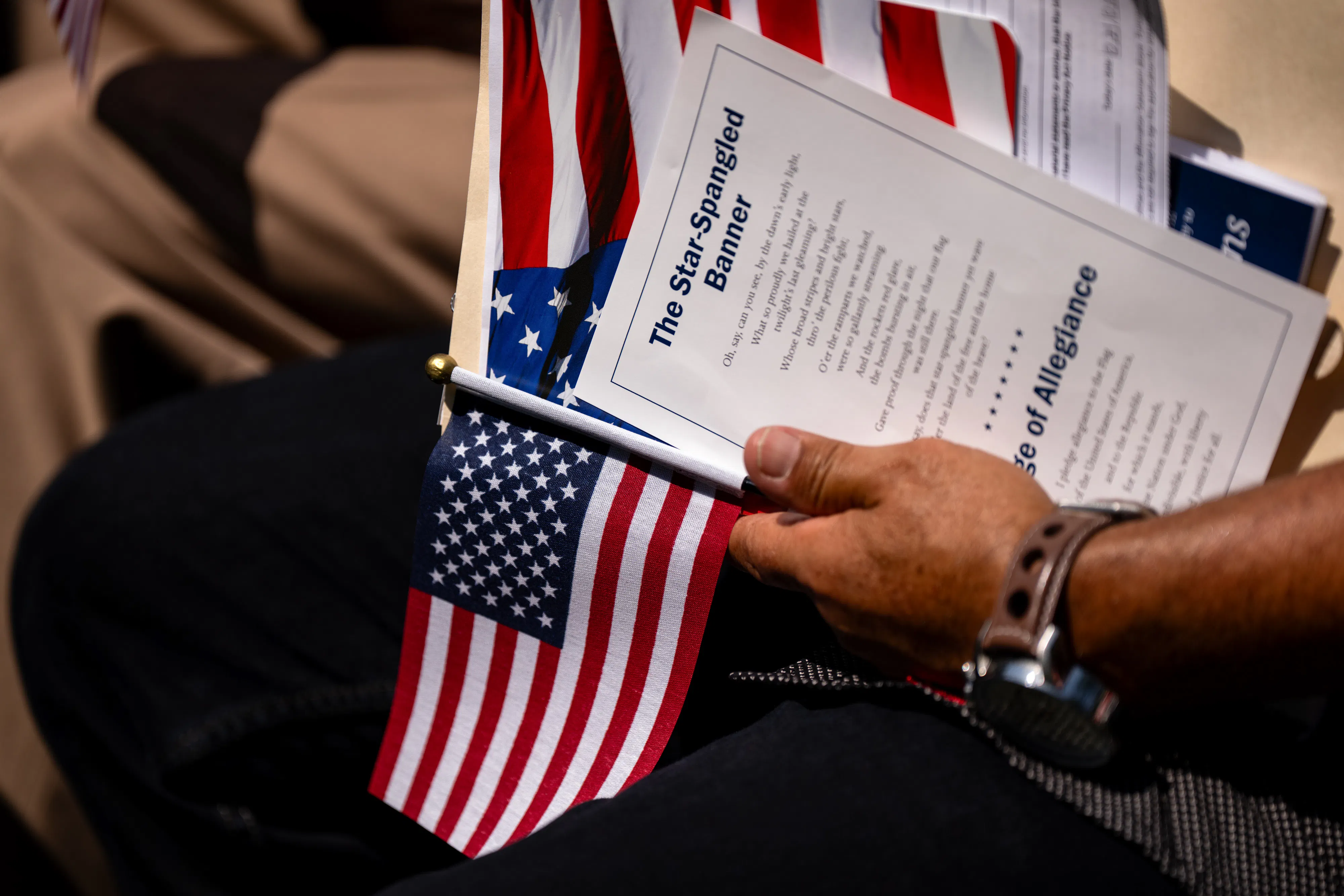 A hand holding an American flag and a printed copy of the Star Spangled Banner lyrics at a citizenship ceremony.