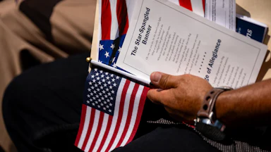 A hand holding an American flag and a printed copy of the Star Spangled Banner lyrics at a citizenship ceremony.