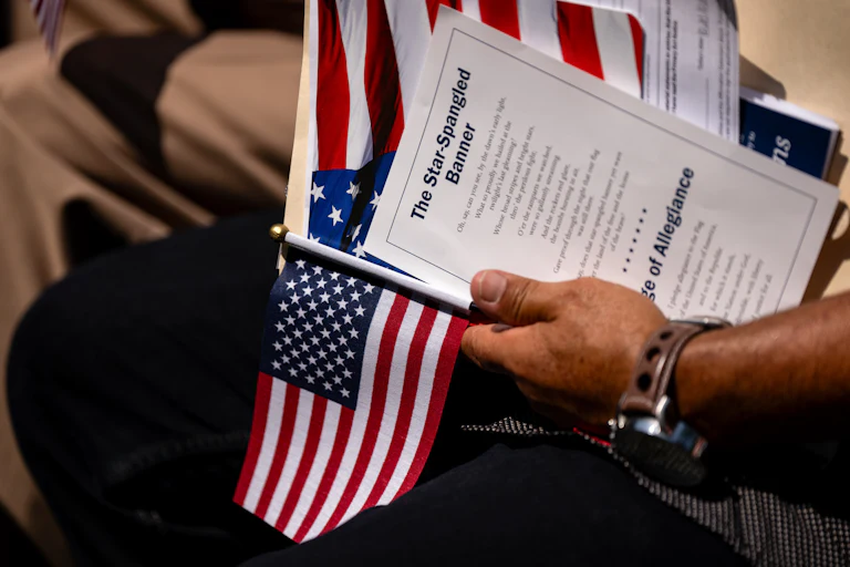 A hand holding an American flag and a printed copy of the Star Spangled Banner lyrics at a citizenship ceremony.