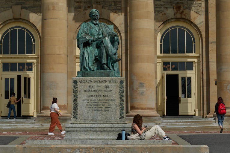 Students sit and walk near a building at Cornell University. In the center of the photo is a green statue of an old white man, Andrew Dickson White, "friend and counsellor of Ezra Cornell."
