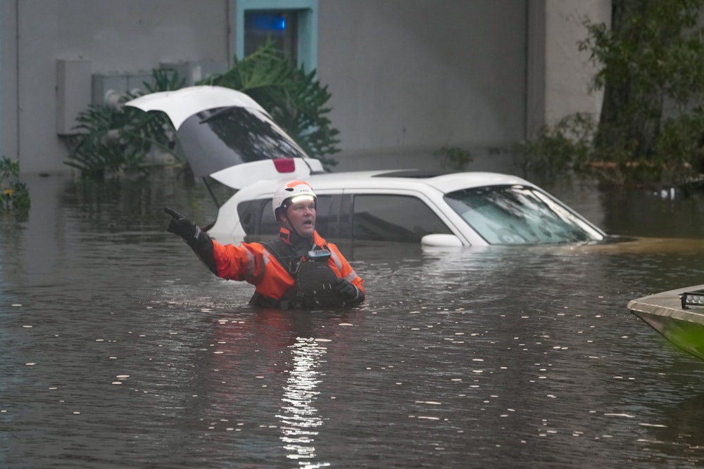 A first responder stands in waist-deep water, with a car and apartment building in the background.