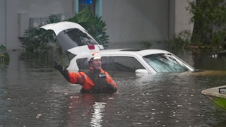 A first responder stands in waist-deep water, with a car and apartment building in the background.