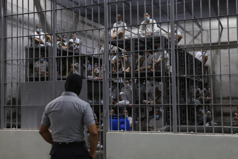 A prison officer guards a cell at CECOT with dozens of men wearing masks sitting on large four-level bunk beds.