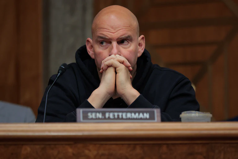 Senator John Fetterman looks to the side while sitting with his hands folded in front of his face
