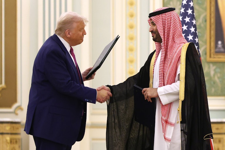 Donald Trump shakes hands with Saudi leader Mohammed bin Salman (wearing traditional attire). Trump holds a black folder in his other hand and he smiles weirdly.