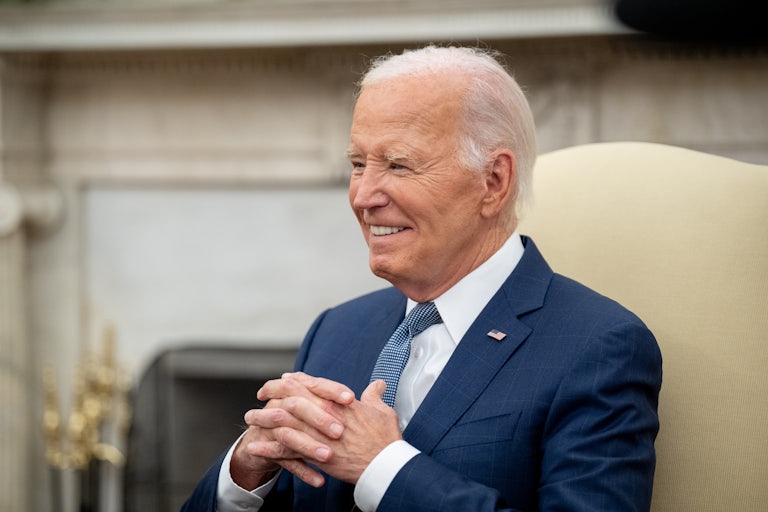 Biden seated, hands intertwined on his chest, smiles