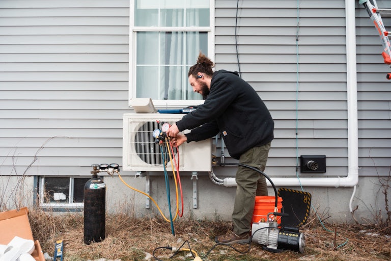 A person bends over a heat pump on the outside of a house, holding wires and gauges.
