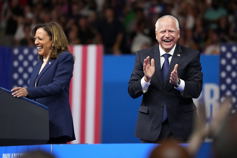 Kamala Harris, standing at a podium, and Tim Walz, clapping, both laugh at a massive campaign rally.