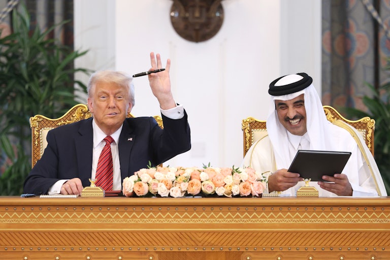 Donald Trump and Emir of Qatar Tamim bin Hamad al Thani sit side by side on gold chairs at a table with flowers. Trump makes a weird half smile and waves with a pen in his hand Hamad al Thani laughs while holding a black folder.