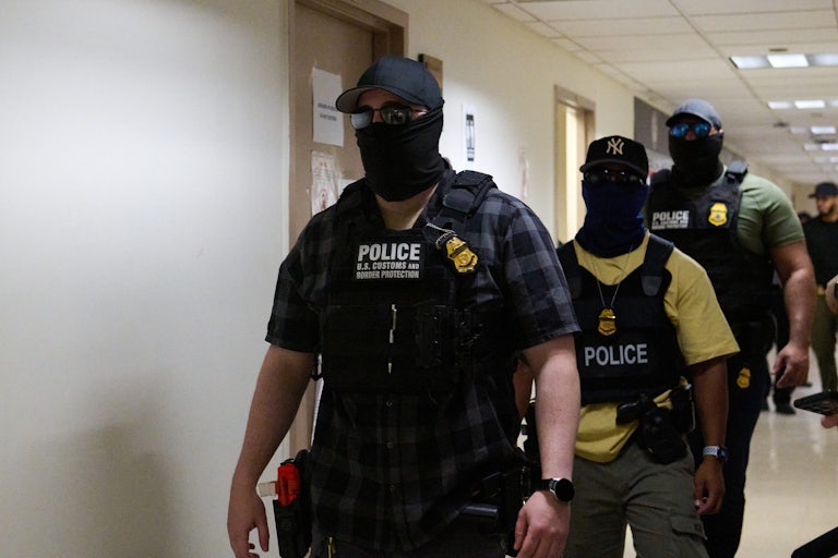 Three men wearing police and border patrol vests walk down a hallway. They are all all wearing sunglasses, caps, and masks to cover their face.