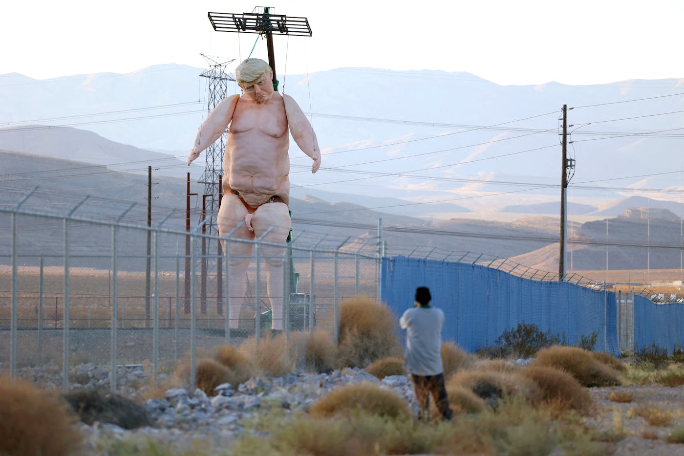 The giant nude Trump statute stands in the fenced-in lot. A person is seen taking a photo of it.