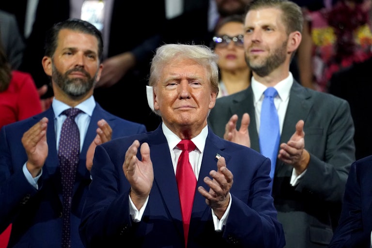 Donald Trump, with a cushion on his right ear, claps and looks straight ahead. Donald Trump Jr. and Eric Trump clap behind him.