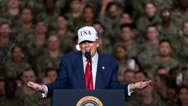 Donald Trump wearing a USA cap shrugs his arms as he speaks to U.S. troops.