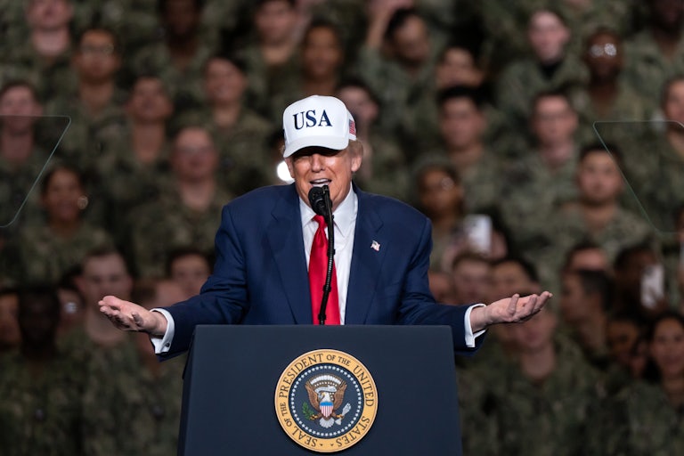 Donald Trump wearing a USA cap shrugs his arms as he speaks to U.S. troops.