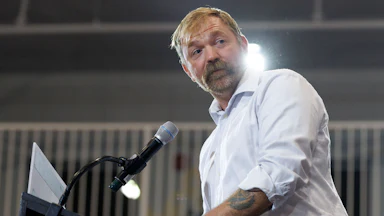 Democratic U.S. Senate candidate Graham Platner stands at a podium, his sleeves rolled up to show his tattoos.