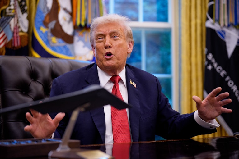 Donald Trump speaks animatedly, hands splayed, while seated at his desk in the Oval Office of the White House.