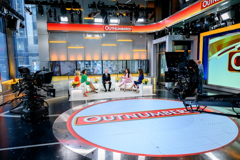 The set of the Fox News Outnumbered. The hosts sit with a guest on white armchairs arranged in a semicircle.