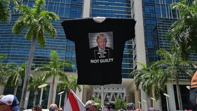 A man holds up a stick with a t-shirt attached to the top that says NOT GUILTY under a photo of Trump.