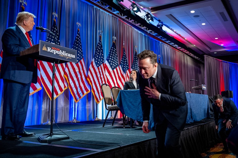 Donald Trump stands at a lectern on the stage while Elon Musk ducks as he tries to walk in front